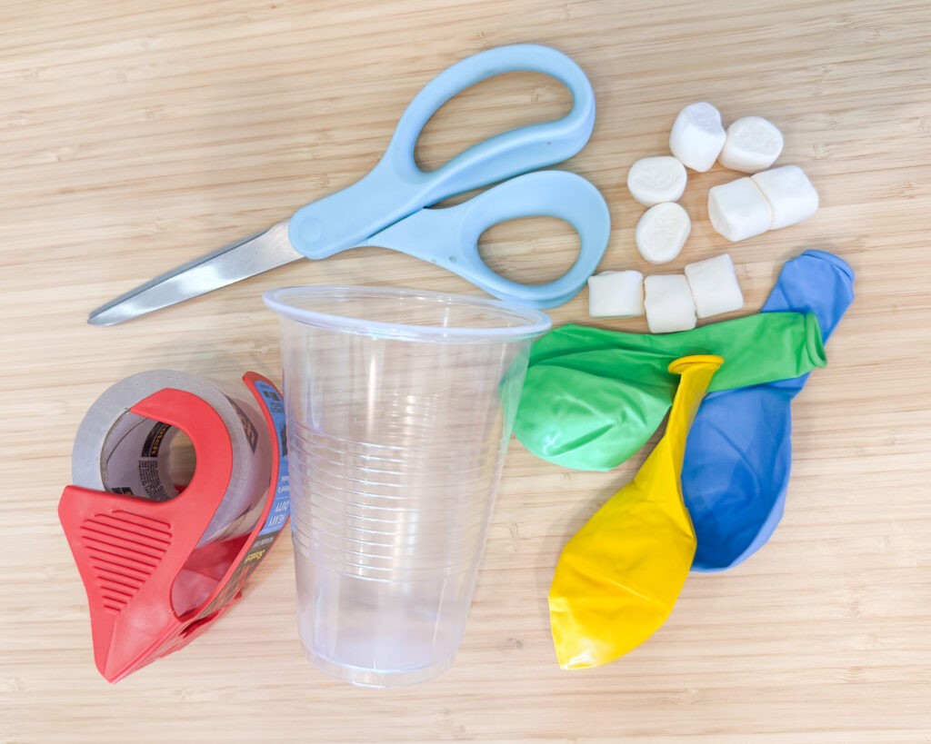 Picture of supplies needed for the project. Pictured are scissors, tape, cup, marshmallows, and balloons.