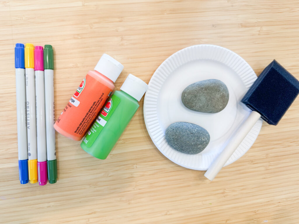 Picture of supplies needed for kindness rock activity. Supplies include rocks, acrylic paint, a paint brush, and paint pends.